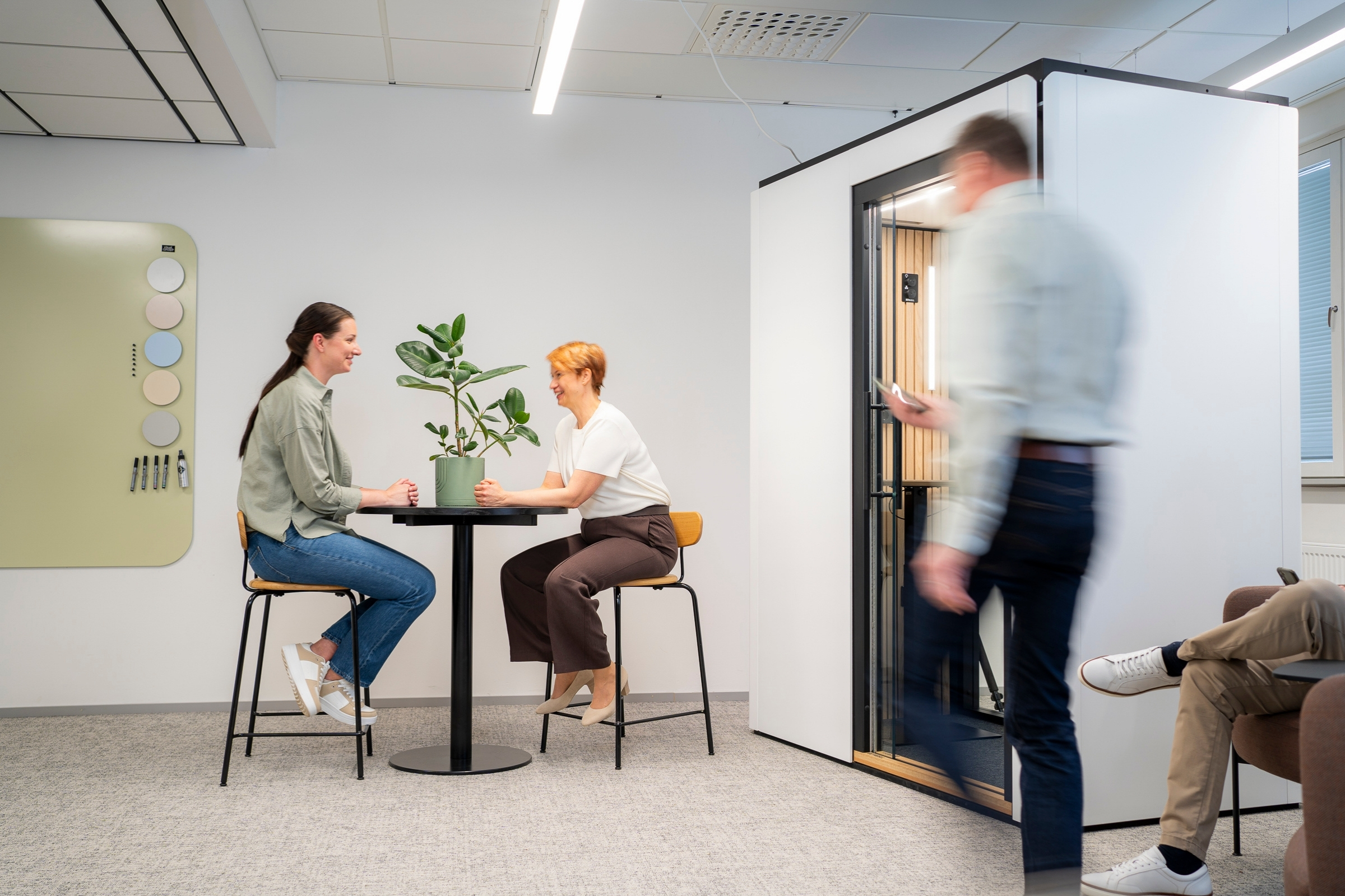 Ihmiset istuvat ja puhuvat taustalla henkilö menossa sisään äänieristettyyn puhelinkoppiin